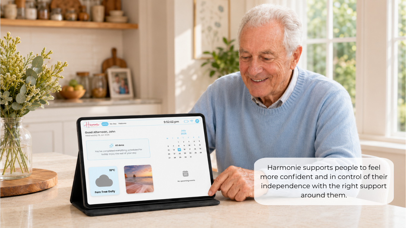 An older man sits at a kitchen table using a tablet device that displays a wellness or calendar app. The scene is brightly lit with natural light from a window in the background, and a text overlay on the right reads, 'Harmony supports people to feel more confident and in control of their independence with the right support around them.