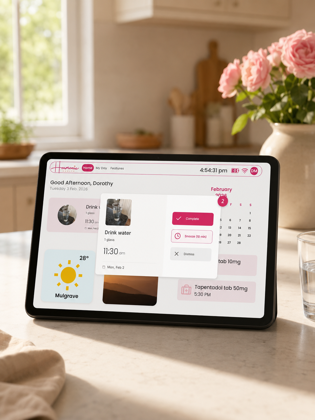 A tablet device on a kitchen counter displaying a digital calendar and planner interface, with a softly blurred kitchen and pink flowers in the background.