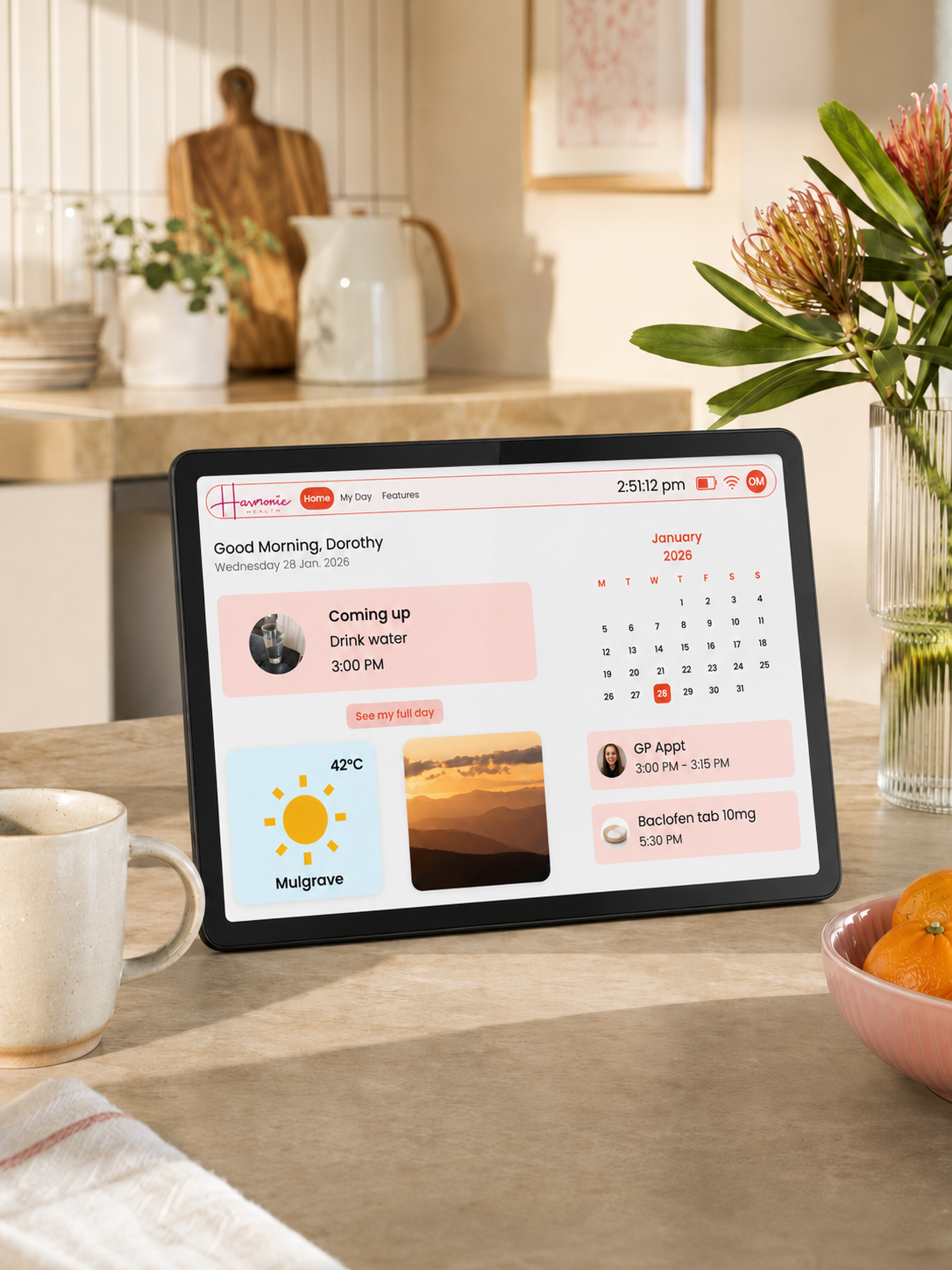 A tablet device displaying a digital calendar and planner interface, placed on a kitchen counter with a mug, flowers, and fruit in a warm, real kitchen environment.