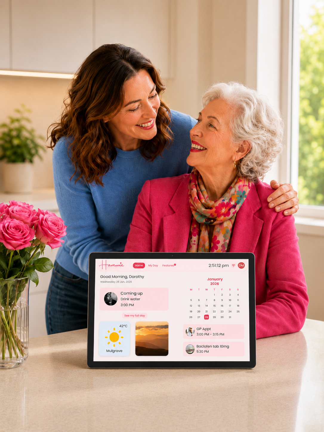 A woman in a blue sweater embraces an elderly woman in a pink blazer seated at a kitchen table. A tablet on the table displays a personalized dashboard interface.