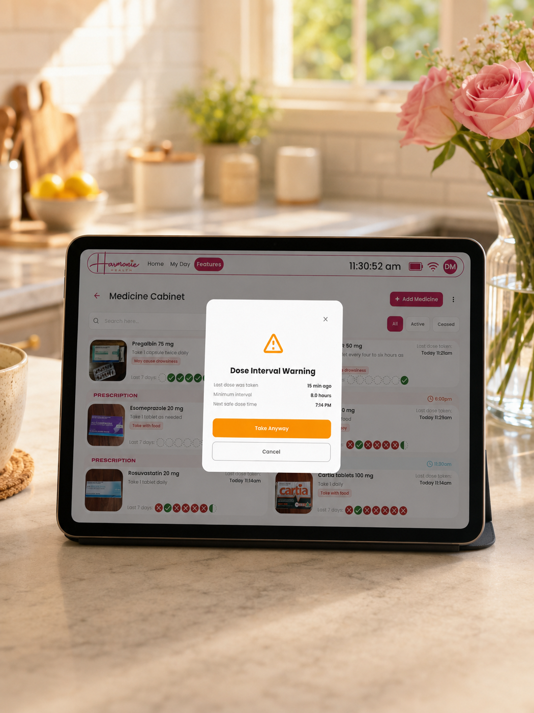 A tablet on a kitchen counter displaying a medicine cabinet app with a dosage warning overlay, set against a softly blurred warm kitchen background with flowers and sunlight.