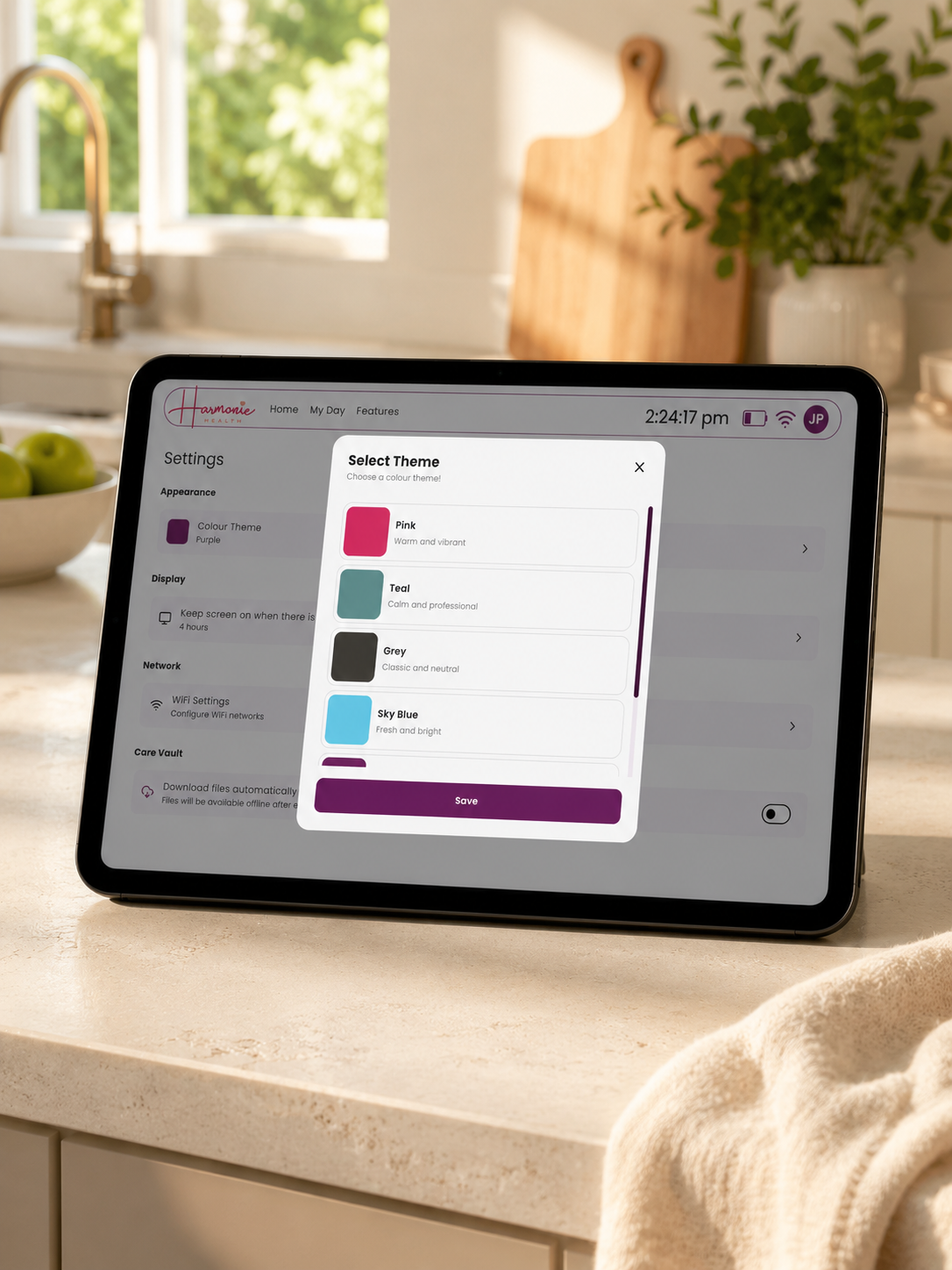 A tablet device on a kitchen counter displaying a settings screen with a pop-up theme selector. The background shows a softly blurred kitchen interior with a sink, faucet, cutting board, and plant.