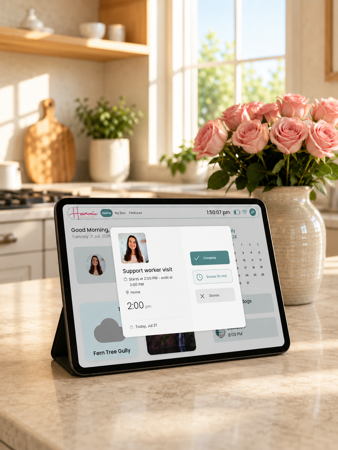 A tablet device on a kitchen counter displaying a digital calendar or scheduling app, with a vase of pink roses and a softly lit kitchen in the background.
