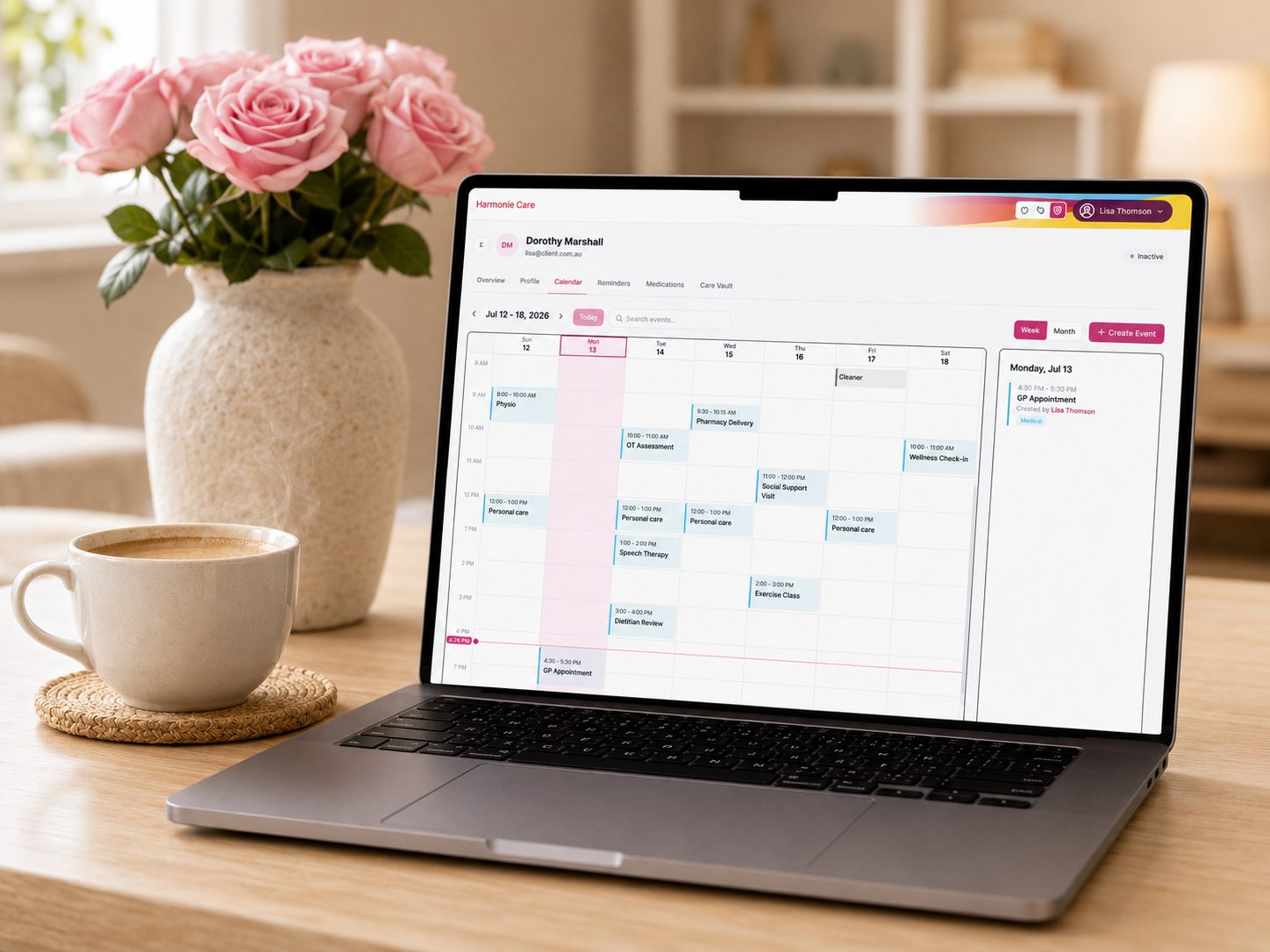 A laptop on a wooden table displaying a digital calendar planner interface, styled with a cup of coffee and a vase of pink roses in a bright, softly lit room.