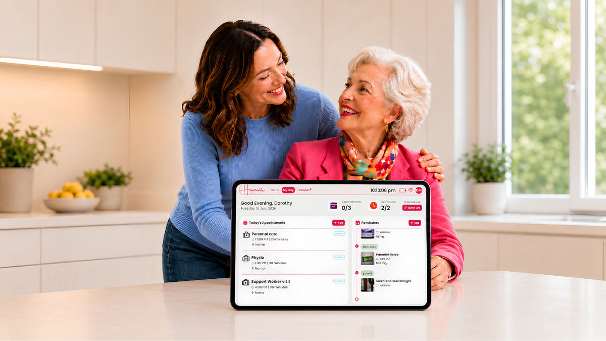 A woman in a blue sweater and an older woman in a pink jacket sit together at a kitchen table, smiling and looking at a tablet screen showing a care plan interface. The scene is set in a bright, modern kitchen with white cabinets and window light.