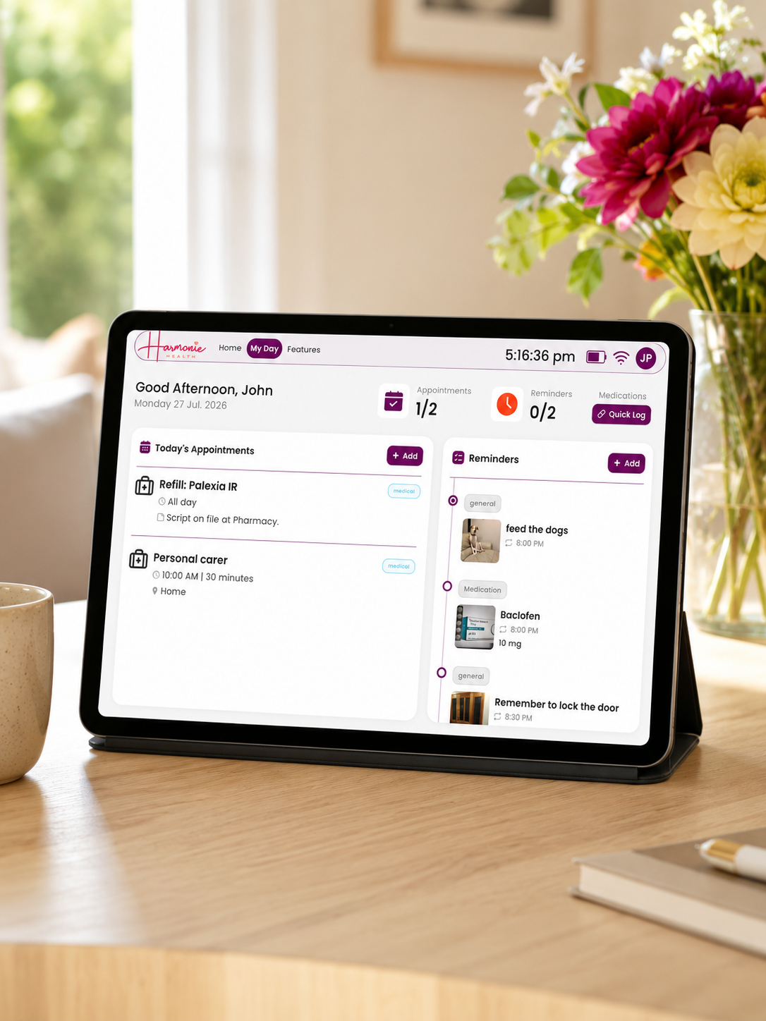 A tablet device on a wooden table displaying a digital calendar or scheduling app interface, with a blurred indoor home background including flowers and a mug.