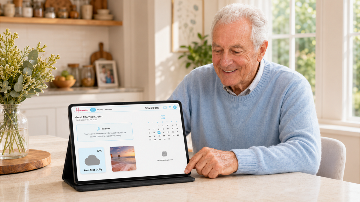 An elderly man with white hair sits at a light-colored wooden table in a bright, modern kitchen, smiling as he uses a tablet device to view a digital weather and calendar app. The tablet is in a black protective case.