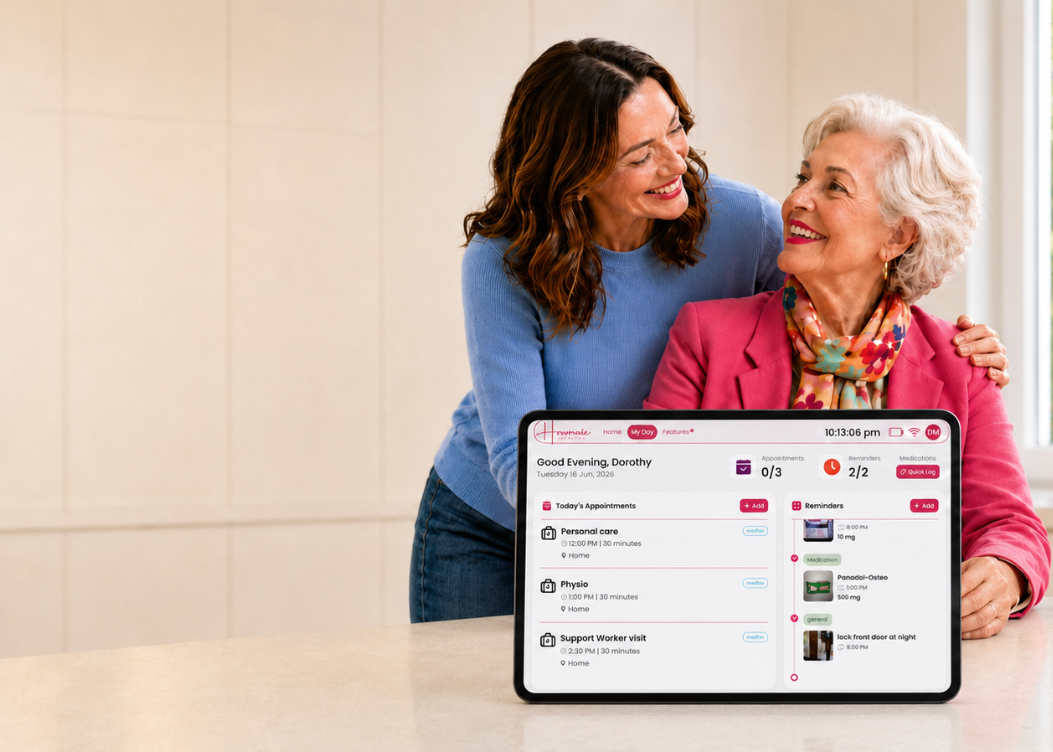 An elderly woman with white hair in a pink blazer sits at a table with a tablet showing a digital care or health dashboard, while a younger woman in a blue sweater leans in from behind. The setting is a bright, modern room with a cream-colored tiled wall and a window.
