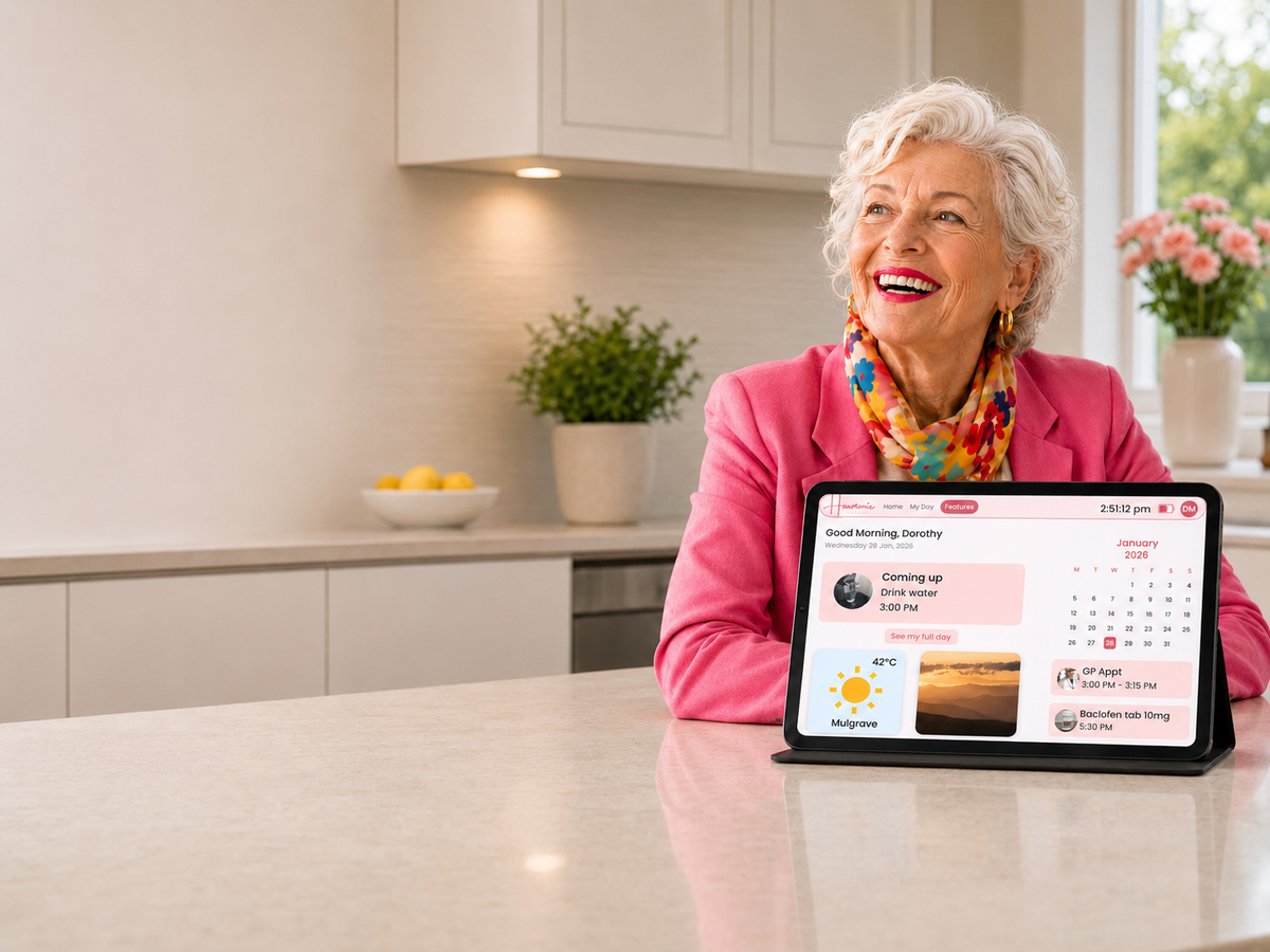An older woman with white hair sits at a kitchen counter smiling, looking at a tablet displaying a digital calendar and weather app. She is wearing a bright pink blazer and a colorful scarf, with a modern kitchen in the background.