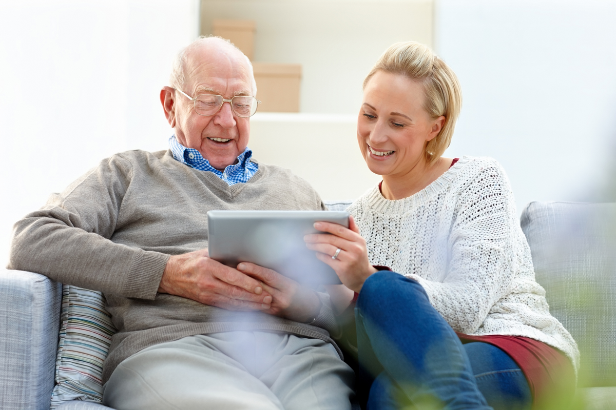 An elderly man and a woman sitting together on a couch, smiling as they look at a tablet device in a bright, modern living room.