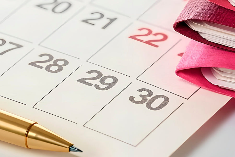 A close-up of a white calendar page with numbered days, a gold pen resting on it, and a stack of pink notebooks on the right side.