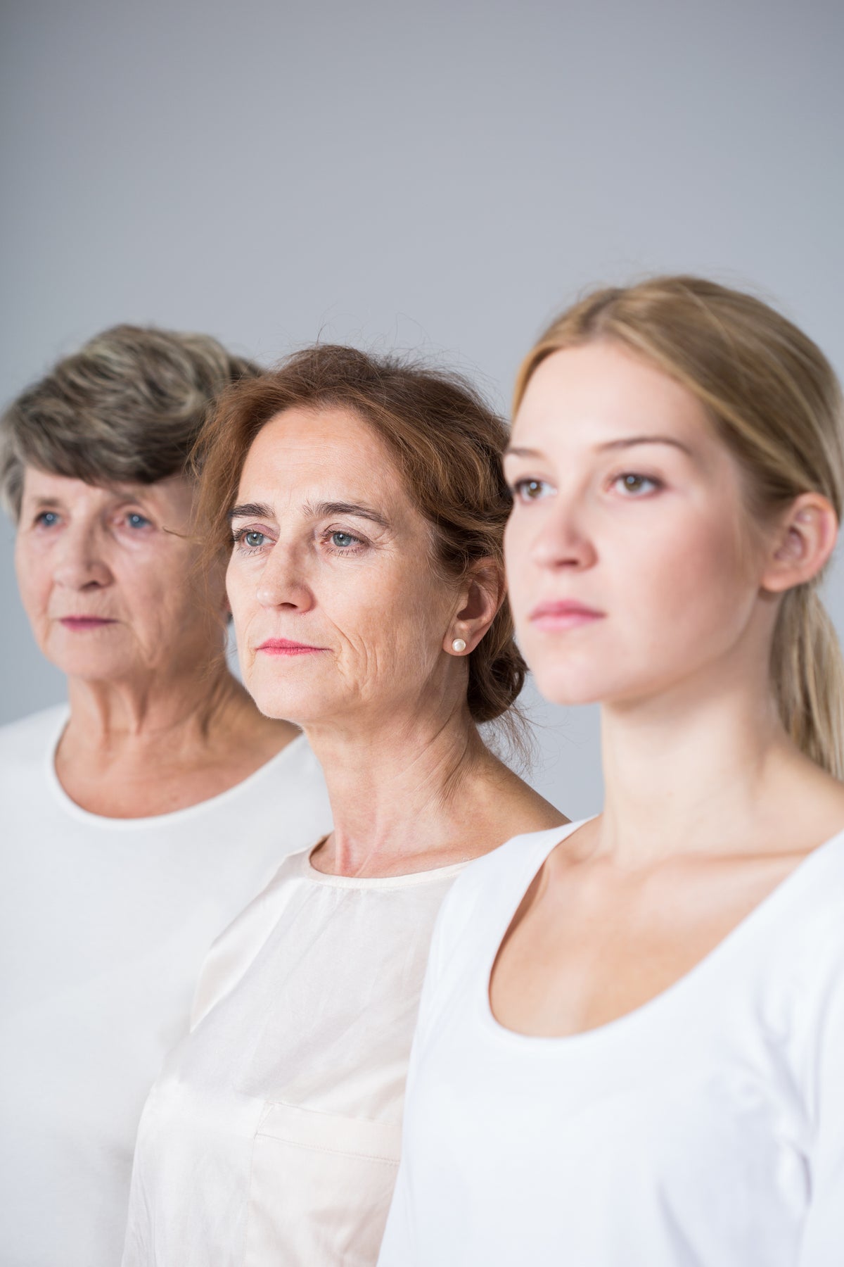 Three women standing side by side in white tops against a plain light gray studio background.