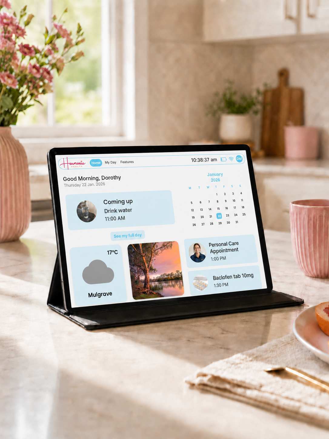 A tablet in a black stand is displayed on a kitchen counter, showing a digital calendar and task interface. The scene is styled with a pink mug, flowers, and a plate in a bright kitchen environment.
